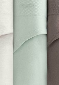 Trois serviettes pliées en blanc doux, vert pâle et brun atténué avec des coutures visibles et une texture subtile.