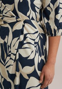 Marineblauwe en crèmekleurige stoffen met een bloemenpatroon en een gestructureerd oppervlak. Het ontwerp heeft grote bladeren en een zijstrikdetail.