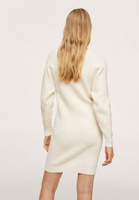 Femme aux longs cheveux blonds portant une robe en maille côtelée crème à manches longues, debout de dos à la caméra.