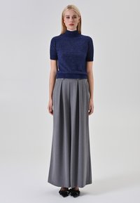 Giovane donna in piedi, che indossa un maglione a maniche corte blu navy con colletto alto, pantaloni grigi a gamba larga, scarpe nere a punta con fiocchi e orecchini ad anello dorati.