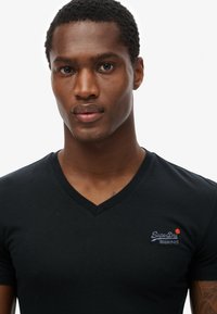 Camiseta negra de cuello en V hecha de algodón, con un pequeño logotipo bordado en el lado izquierdo del pecho. Textura suave, diseño ajustado.