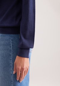 Granatowy sweter dziany z długimi rękawami, z prążkowanymi mankietami, w połączeniu z jasnoniebieskimi dżinsami. Gładka faktura, dopasowany krój, widoczne detale szycia.