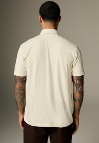 Kortærmet beige skjorte med et geometrisk mønster, blød tekstur, krave med spread og subtile designdetaljer. Mørke bukser synlige.