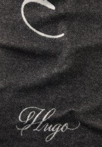Tissu gris foncé avec du texte en cursive blanc "Hugo" et une ligne courbe partielle blanche au-dessus du texte.