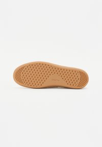 Suola di una scarpa caratterizzata da una superficie in gomma testurizzata di colore marrone chiaro, con un motivo a diamante e logo impresso, forma piatta e leggermente curvata.