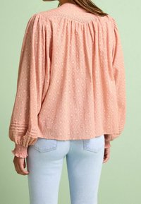 Blusa di colore pesca con texture, maniche lunghe e voluminose e arricciature sui polsini. Yoke posteriore a forma di V con cuciture decorative. Indossata sopra jeans azzurri.