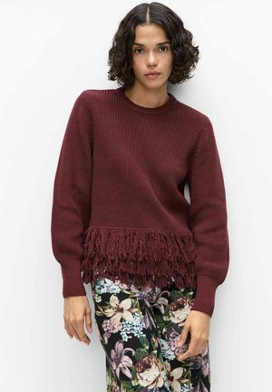 Kvinde med kort krøllet hår iført en vinrød strikket sweater med frynser og bukser med blomstermønster, der står foran en hvid baggrund.