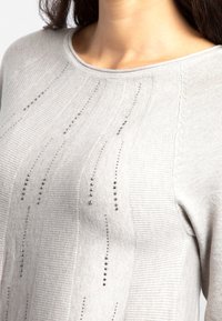 Femme portant un pull en tricot gris clair avec des rangées verticales de petits clous métalliques, un col rond et des motifs ribbés texturés.