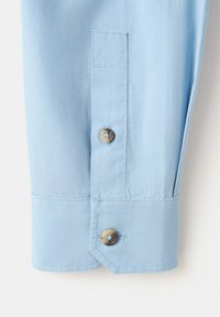 Manche de chemise bleu clair avec poignet boutonné et deux boutons marron, montrant une couture détaillée sur un tissu lisse.