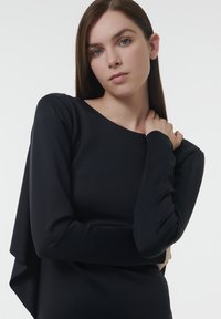 Zwarte lange mouw top van een gladde, rekbare stof; heeft een asymmetrisch ontwerp met een gedrapeerde laag aan één kant, wat een getailleerde silhoeët biedt.