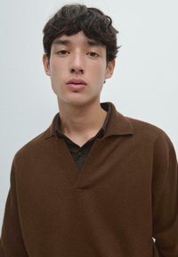 Brauner Strickpullover mit V-Ausschnitt und geripptem Kragen, über einem dunklen karierten Hemd getragen. Glatte Textur, lockere Passform.