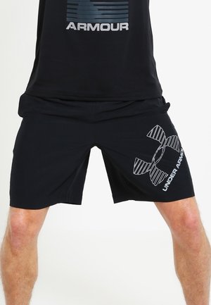 Mand iført sorte Under Armour-shorts og sort Under Armour-t-shirt, stående med benene spredt mod en ensfarvet baggrund.
