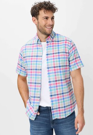 Mann mit lockigem Haar, der ein kurzärmeliges, pastellkarierte Hemd über einem weißen T-Shirt und blauen Jeans trägt, lächelnd und nach rechts schauend.