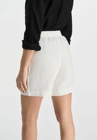 Białe lniane szorty z elastycznym paskiem, bocznymi kieszeniami i luźnym krojem, pokazane z tyłu, w połączeniu z czarną koszulą z długim rękawem.