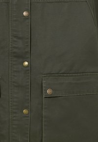Veste olive en vert avec une texture lisse, des boutons-pression et une poche avant fermée par un seul bouton. Design simple et détails pratiques.