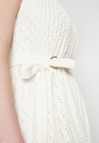 Robe blanche présentant un motif texturé, taille cintrée avec une ceinture à nouer, et un nœud décoratif au niveau de la ceinture.