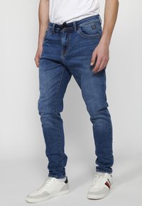 Jeans de denim azul ajustados con un acabado ligeramente descolorido, presentan una cinturilla con cordón, cinco bolsillos y detalles de costura sutiles.
