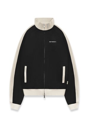 Mann trägt eine schwarz-beige Jacke mit Reißverschluss und passende Hose mit dem Text "RETERNITY", steht vor einem einfarbigen hellen Hintergrund.