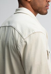 Baardige man in een lichtbeige blouse met kraag, gezien vanaf de schouder en in zijaanzicht tegen een neutrale achtergrond.