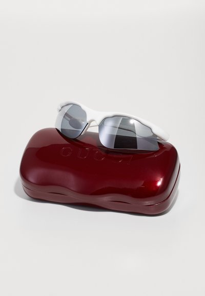 Occhiali da sole bianchi dal design elegante e con lenti scure poggiati su un astuccio rosso lucido con il logo in rilievo "GUCCI."