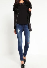 Chaqueta con cuello de piel sintética negra sobre una blusa negra, combinada con vaqueros ajustados azules desgastados y zapatos planos negros. Textura lisa y corte entallado.