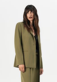 Olive green blazer with a tailored fit, single button closure, lapel collar, and side pockets, paired with a black top; smooth texture.