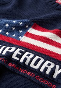 Pull en maille bleu marine avec un motif du drapeau américain en bleu, rouge et blanc. Le texte "SUPERDRY" est affiché en évidence en broderie blanche et rouge.