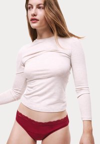 Långärmad, ljusbeige topp med draperad halsringning, kombinerad med röda underkläder med spetsdetaljer vid midjan.