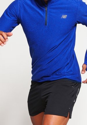 Persoon die een blauw atletisch shirt met lange mouwen en een rits draagt en zwarte hardloopshorts, vastgelegd in beweging tegen een effen achtergrond.