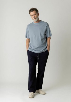 Homme à la barbe grise portant une chemise à manches courtes bleu clair, un pantalon bleu marine foncé, des baskets blanches, debout les mains dans les poches sur un fond neutre.