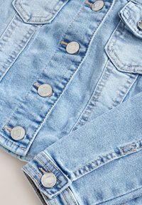 Veste en denim bleu clair avec boutons-pression métalliques, poche poitrine et coutures visibles sur les manches et le panneau avant.
