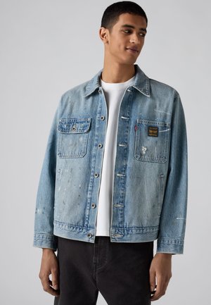 Junger Mann trägt eine hellblaue Jeansjacke mit Fronttaschen, ein weißes T-Shirt und schwarze Hose, schaut nach links.