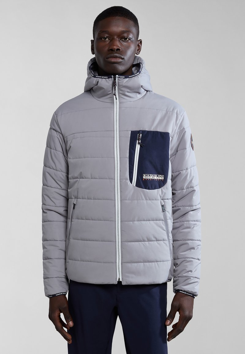 Chaqueta acolchada gris con un bolsillo azul marino con cremallera, cuello alto y puños elásticos. Presenta cremallera de contraste y un logo en el bolsillo.
