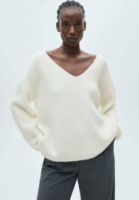 Cremig farbener Strickpullover mit V-Ausschnitt, lockerer Passform, überschnittenen Schultern und gerippter Textur, kombiniert mit grauen Nadelstreifenhosen.