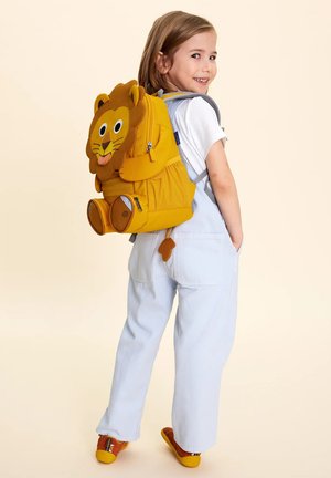 Young child smiling and looking back, wearing white overalls, orange shoes, and a yellow lion-themed backpack.
