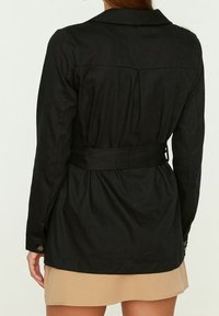 Giacca nera con cintura, maniche lunghe, colletto e retro sagomato. Presenta polsini con bottoni e un tessuto testurizzato.