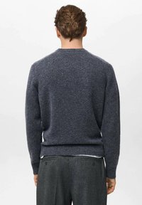 Uomo con capelli corti castani di spalle, che indossa un maglione grigio scuro e pantaloni grigi abbinati, su uno sfondo bianco semplice.