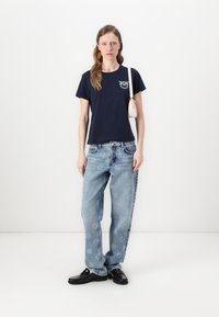 T-shirt cropped blu navy con design ricamato, abbinato a jeans ampi di colore azzurro chiaro con motivi floreali e scarpe nere. Borsa a spalla rosa.