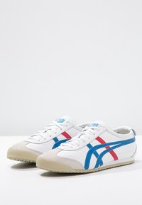 Paire de baskets blanches avec des rayures bleues et rouges, bouts beiges et semelles beige clair sur une surface blanche.