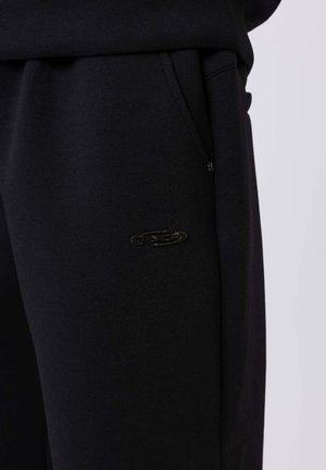 Pantalones deportivos negros hechos de un material suave, con un bolsillo lateral, cintura elástica y un pequeño logo negro en el muslo.