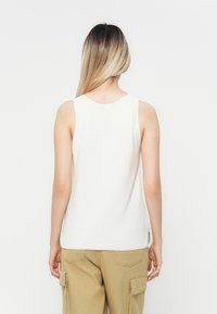White sleeveless top with a textured finish, round neckline, and side slits, paired with khaki cargo pants.