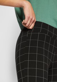 Svarte rutete leggings med bred livlinje, som har hvite rutenettlinjer og et mykt, teksturert materiale. Hånd plassert på livlinjen.