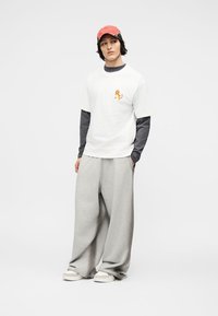 Νεαρός που φοράει κόκκινο καπέλο, λευκό μπλουζάκι με μικρό καρτούν χαρακτήρα, από πάνω σκούρο μακρυμάνικο πουκάμισο και φαρδιά γκρι παντελόνια.