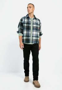 Geruite flanellen overhemd in marineblauwe, witte en groene tinten, gedragen over een groene crew neck shirt. Gecombineerd met zwarte broeken en beige schoenen.