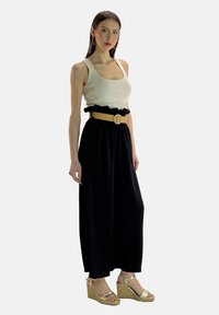 Cremefarbener Tanktop, schwarze Hosen mit weitem Bein und Rüschensaum am Bund, gewebter beiger Gürtel mit runder Schnalle und goldene Keilsandalen. Schlichter Hintergrund.