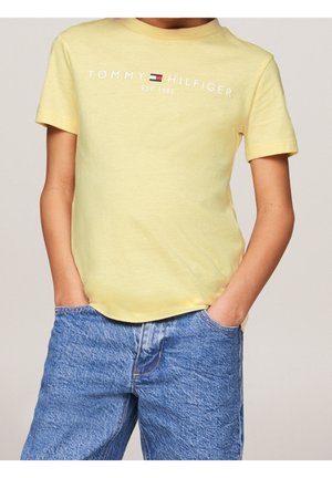 Dziecko w żółtej koszulce Tommy Hilfiger z logo i niebieskich dżinsach, ręce w kieszeniach, na jednolitym tle.