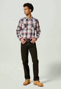 Camisa xadrez vermelha, branca e azul com dois bolsos no peito, calças pretas e botas de salto beje, de pé contra um fundo neutro.