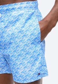 Μπλε μαγιό βερμούδα με λευκό μοτίβο γραφής, ελαστική μέση, πλαϊνή τσέπη και μικρό λογότυπο κόκκινου, λευκού και μαύρου χρώματος κοντά στο τελείωμα.