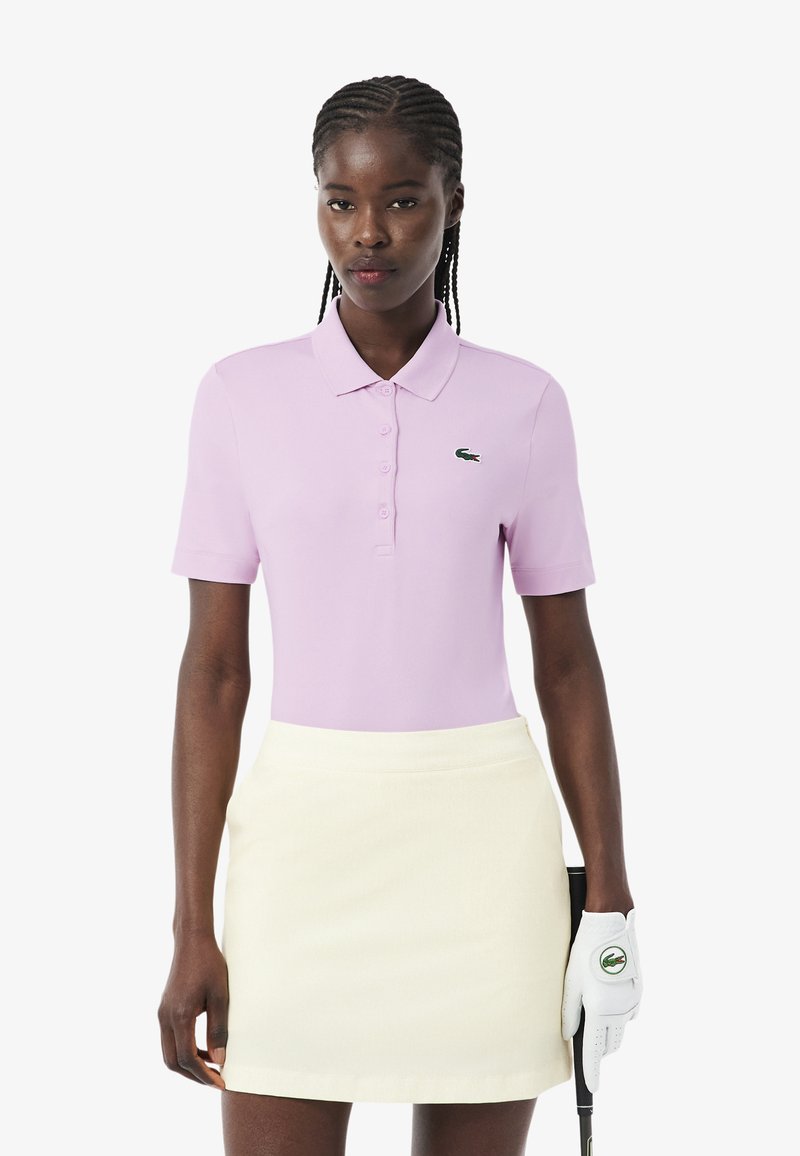 Mujer con cabello trenzado, vestida con un polo lila claro, falda crema y guante de golf, sosteniendo un palo de golf sobre un fondo blanco.