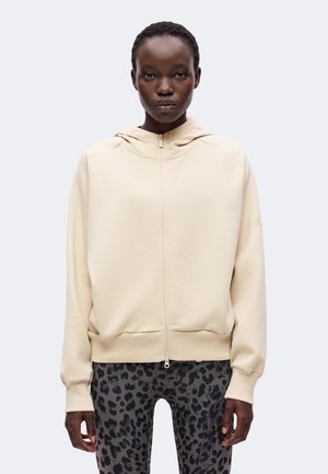 Personne portant un sweat à capuche beige zippé et des leggings noirs et gris à imprimé léopard, se tenant devant un fond clair uni.